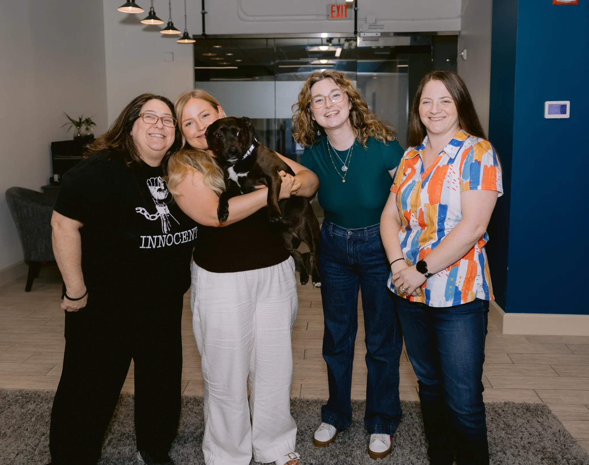 Group of four women posing with a black dog in a modern office lobby, smiling at the camera.