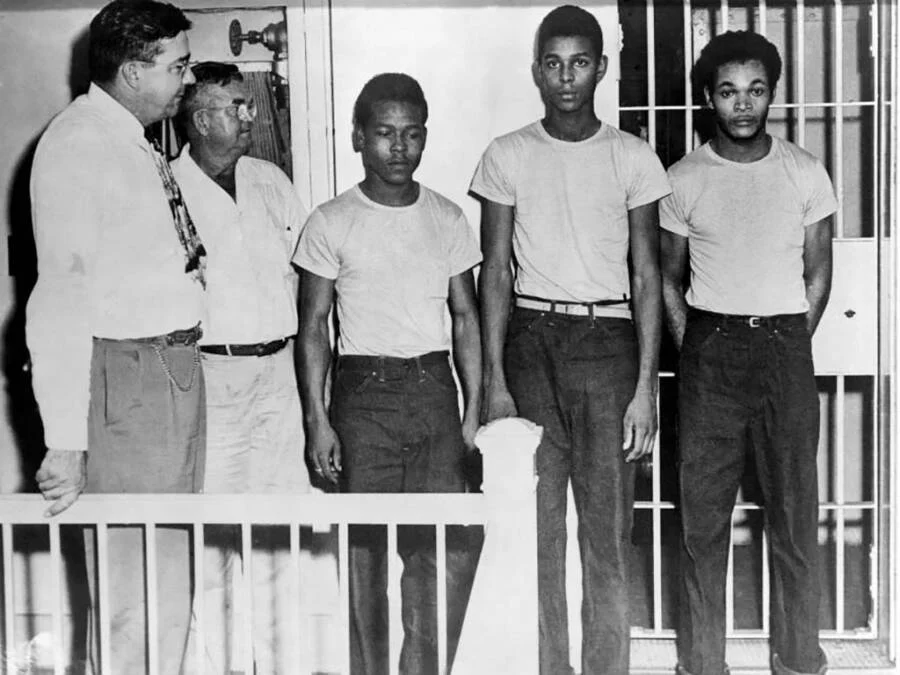 Three young Black men stand in a line in front of a barred cell, flanked by two older men in civilian clothes nearby.