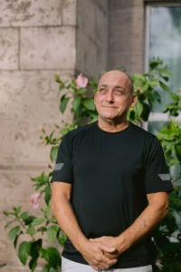 - Innocence Project of Florida Smiling, bald man in a black T-shirt stands outdoors near a stone wall and green plants.
